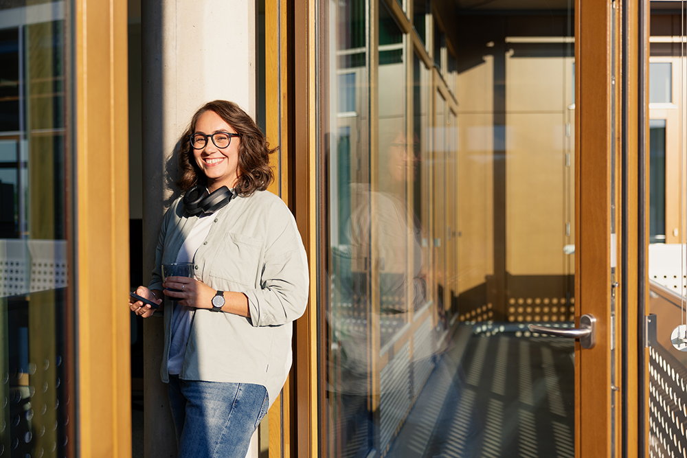 Junge Frau mir Headset steht vor offener Bürotüre und hat Smartphone in der Hand