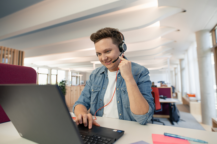 Auszubildender mit Headset vor Laptop in Open-Space-Büro im dm-dalogicum