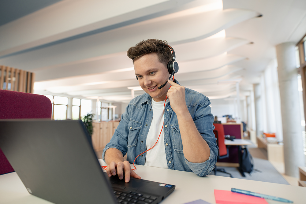 Auszubildender mit Headset vor Laptop in Open-Space-Büro im dm-dalogicum