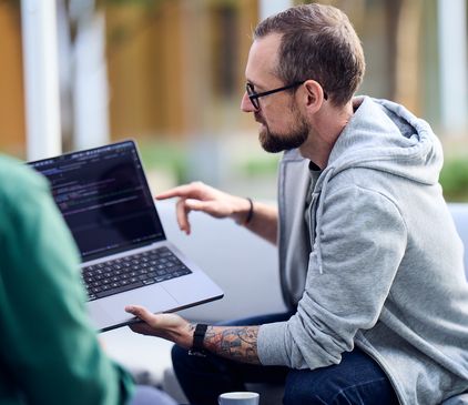 Ein Mann in Kapuzenpulli sitzt draußen und zeigt seinem Kollegen Code auf seinem Laptop
