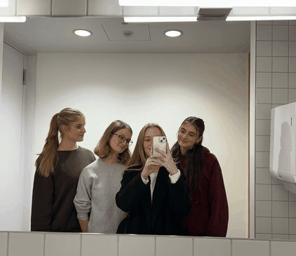 Vier lächelnde junge duale Studentinnen machen gemeinsam ein Spiegelselfie in einem gefliesten Bad