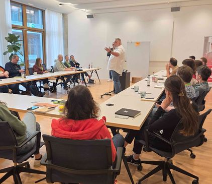 In einem hellen Besprechungsraum im dm-dialogicum sitzt eine Gruppe von Agile Lean Karlsruhe-Teilnehmenden an U-förmig angeordneten Tischen und hört einem stehenden Vortragenden zu.
