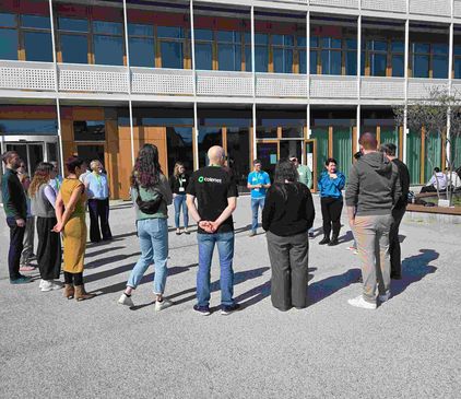 Eine Gruppe von etwa zwanzig Erwachsenen steht draußen im Kreis vor dem dm-dialogicum und hört einer Person in blauer Jacke zu.