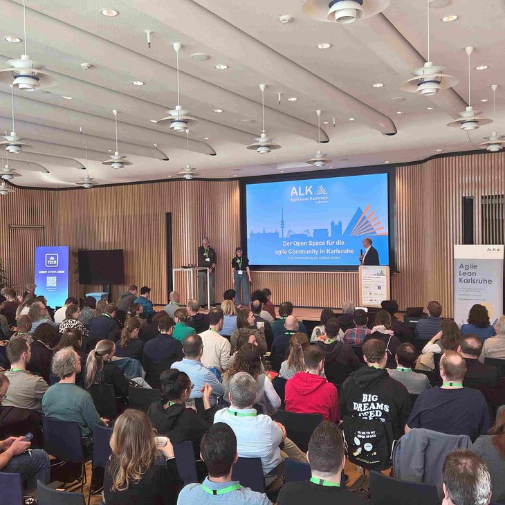 Voller Besprechungsraum im dm-dialogicum mit zahlreichen sitzenden Teilnehmenden, die zwei Vortragenden auf einer Bühne zuhören, die vor einer großen Leinwand mit der Aufschrift „ALK Agile Lean Karlsruhe – Der Open Space für die agile Community in Karlsruhe“ stehen.