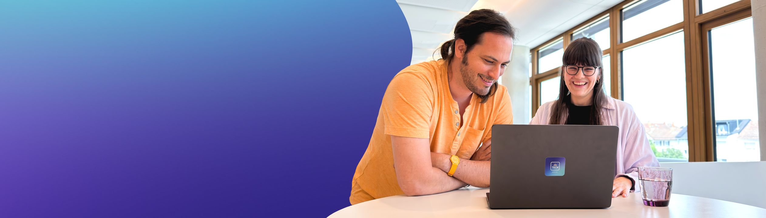 Zwei lächelnde Mitarbeitende sitzen an einem Tisch vor einem Laptop mit dmTECH-Logo. Der Mann links trägt ein orangefarbenes Hemd und eine gelbe Uhr, die Frau rechts trägt ein rosa Hemd und eine Brille. Beide schauen auf den Bildschirm und wirken engagiert und zufrieden bei der Zusammenarbeit.