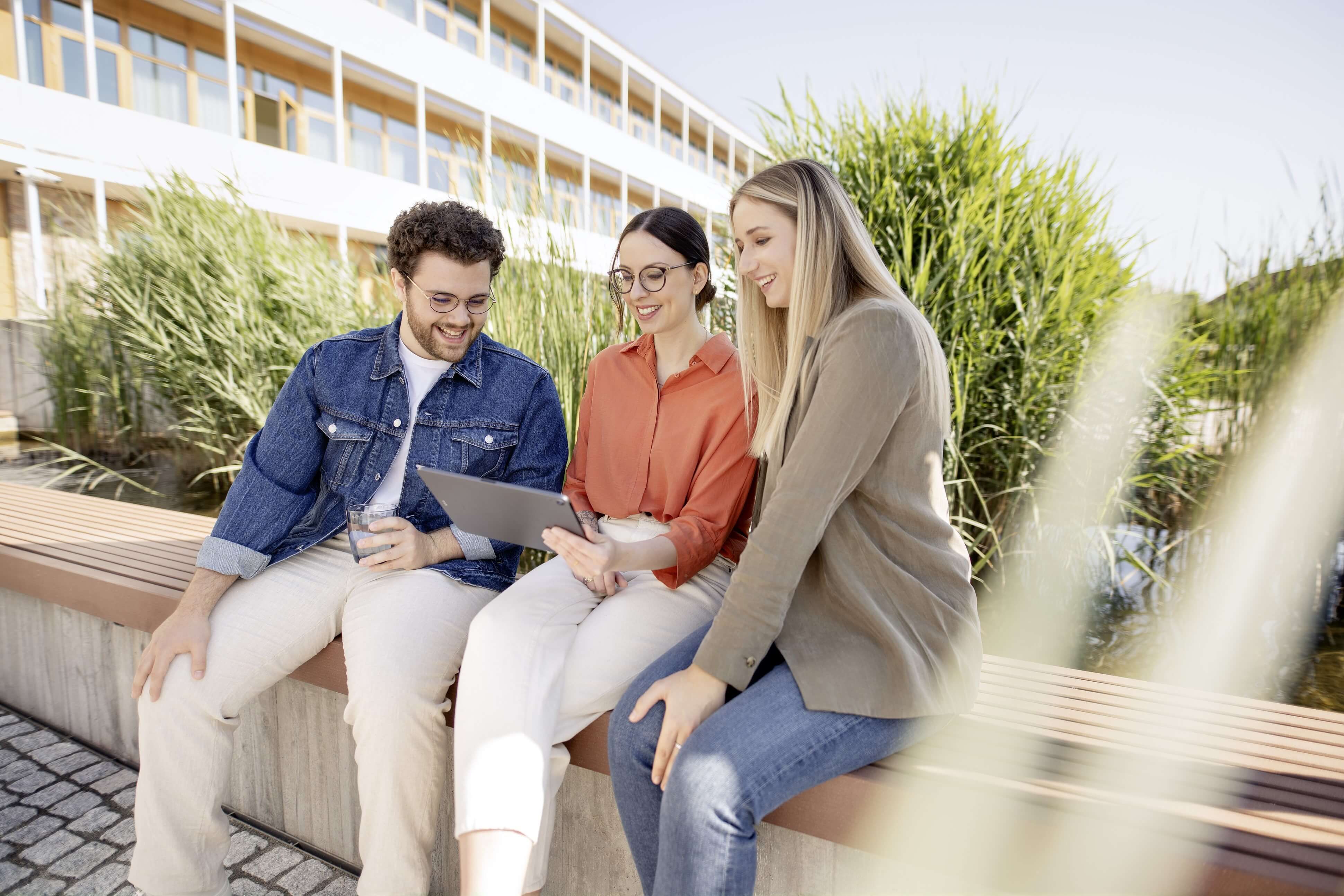 Ein junger IT-Kollege und zwei junge IT-Kolleginnen sitzen draußen auf der Terrasse vor dem Mitarbeiterrestaurant und schauen gemeinsam auf ein Tablet