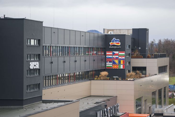 Moderne Bürogebäude des dialogicums in Wals bei Salzburg mit dunkelgrauer Fassade und beigen Elementen, dm dialogicum Schriftzug mit Logo oben, große Flaggentafel mit Flaggen verschiedener europäischer Länder, darunter Dachterrasse mit getrockneten Gräsern, im Hintergrund weitere Gebäude und bewaldete Hügel