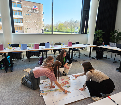 Vier Schülerinnen hocken um ein großes Plakat herum und sortieren ausgeschnittene Fotos von dm-Produkten in ein aufgezeichnetes Regalschema ein. Im Hintergrund ist das dm-dialogicum in Karlsruhe zu sehen.