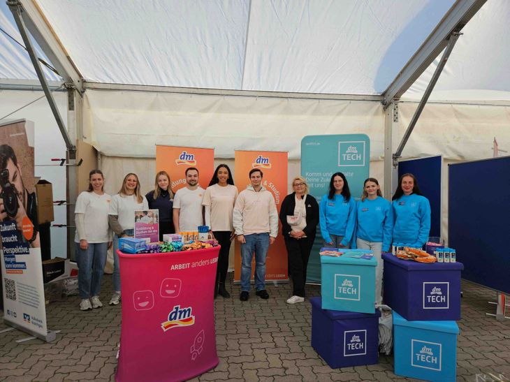 In einem Messezelt auf der Ausbildungsplattform Stutensee stehen dm- und dmTECH-Kollegen vor Roll-ups zu Ausbildung und Studium bei dm und dmTECH, vor ihnen stehen ein pinker dm-Infotresen mit Give-aways sowie mehrere lila und blaue Würfel mit dem dmTECH-Logo, auf denen weitere Produkte und Infomaterial liegen.
