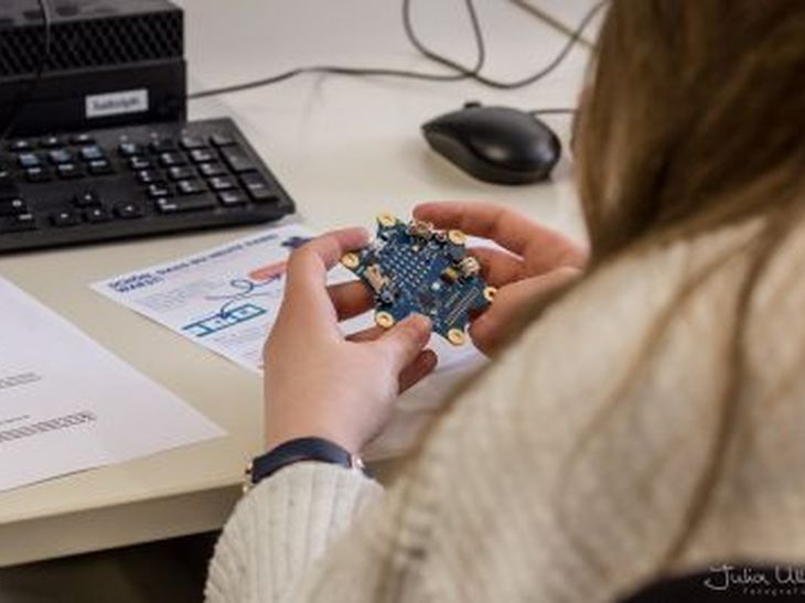 Schülerin sitzt vor einer Tastatur und einer PC-Maus und hält eine Platine in der Hand