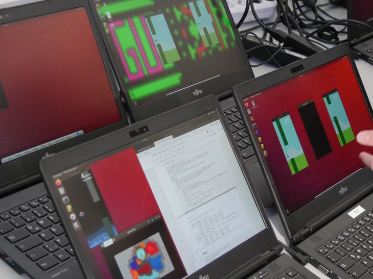 Mehrere Laptops mit Code und im rechten Bildrand eine Hand, die auf das Dargestellt deutet.