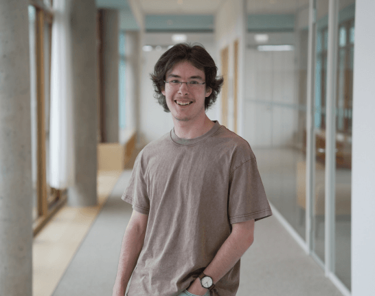 Ein junger Auszubildender Fachinformatiker mit Brille und lockigem dunklem Haar steht lächelnd in einem hellen Flur und trägt ein beigefarbenes T-Shirt.