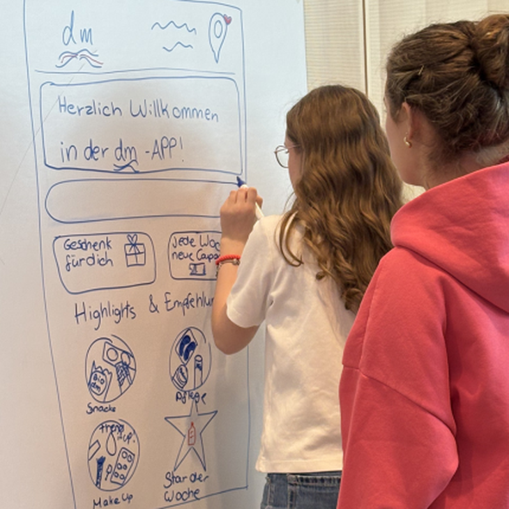 Zwei Schülerinnen des Girls Day bei dmTECH stehen mit dem Rücken zur Kamera und gestalten auf einem Whiteboard neue Vorschläge für die dmApp.
