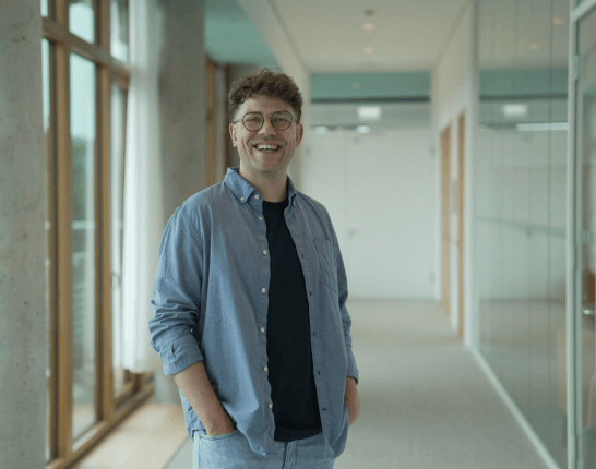 Ein Student im Programm StudiumPLUS mit Brille und lockigen Haaren steht entspannt in einem hellen Flur mit großen Fenstern und Glaswänden und lächelt.