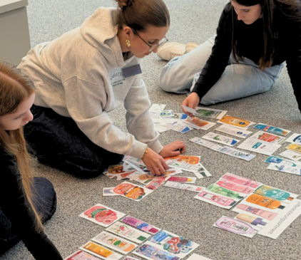 Drei Schülerinnen beim Girls Day sitzen auf dem Boden und sortieren verschiedene auf Papier gedruckte dm-Produkte ihren Produktkategorien zu.