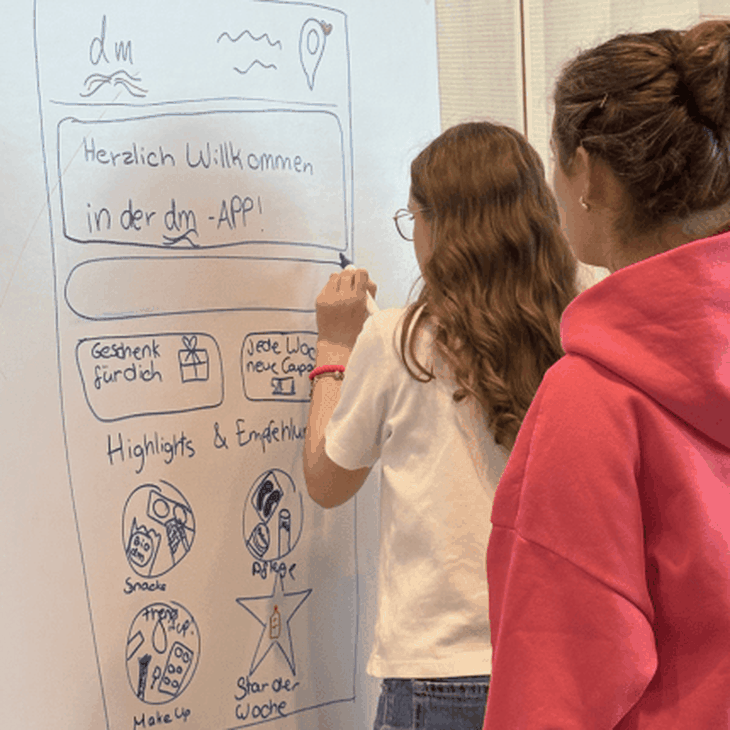Zwei Schülerinnen des Girls' Day bei dmTECH stehen mit dem Rücken zur Kamera und gestalten auf einem Whiteboard neue Vorschläge für die dmApp.