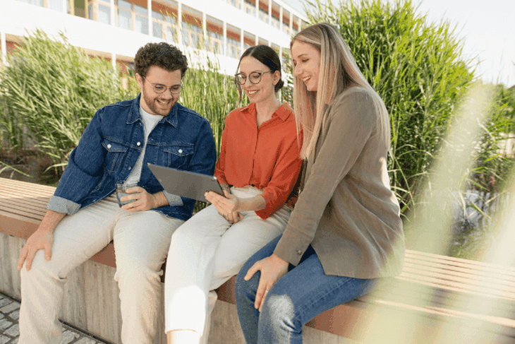 Ein junger IT-Kollege und zwei junge IT-Kolleginnen sitzen draußen auf der Terrasse vor dem Mitarbeiterrestaurant und schauen gemeinsam auf ein Tablet