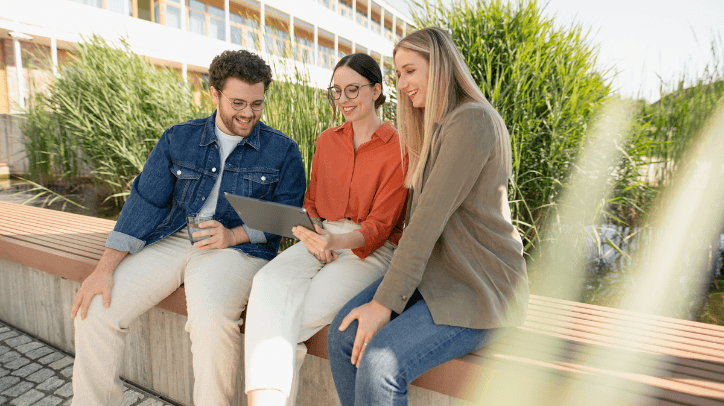 Ein junger IT-Kollege und zwei junge IT-Kolleginnen sitzen draußen auf der Terrasse vor dem Mitarbeiterrestaurant und schauen gemeinsam auf ein Tablet