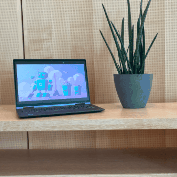 Holzoberfläche mit aufgeklapptem Laptop mit bunter Roboter-Illustration, daneben eine grüne Zimmerpflanze im grauen Topf und ein rosafarbenes Sitzkissen