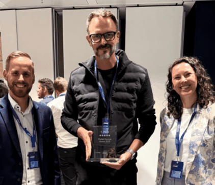 Drei dmTECH Mitarbeitende stehen in einem Veranstaltungsraum, der Mitarbeiter in der Mitte hält die gläserne Trophäe des „Digital Service Award 2025“ für die dm-App in den Händen