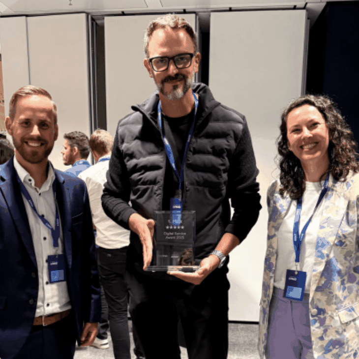 Drei dmTECH Mitarbeitende stehen in einem Veranstaltungsraum, der Mitarbeiter in der Mitte hält die gläserne Trophäe des „Digital Service Award 2025“ für die dm-App in den Händen