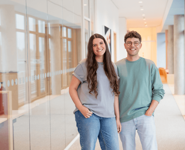 Eine duale Studentin und ein dualer Student stehen im Flur des dm-Unternehmenssitzes in Karlsruhe und lächeln in die Kamera.
