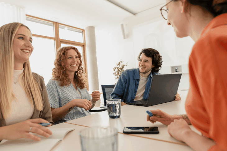 Drei junge IT-Mitarbeiterinnen und ein junger IT-Mitarbeiter sitzen in einem Meetingraum im dm-dialogicum und lachen gemeinsam
