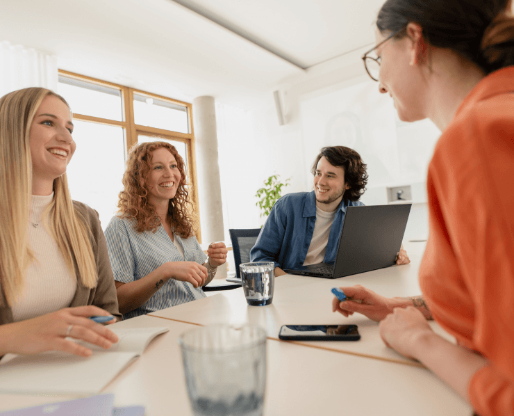 Drei junge IT-Mitarbeiterinnen und ein junger IT-Mitarbeiter sitzen in einem Meetingraum im dm-dialogicum und lachen gemeinsam