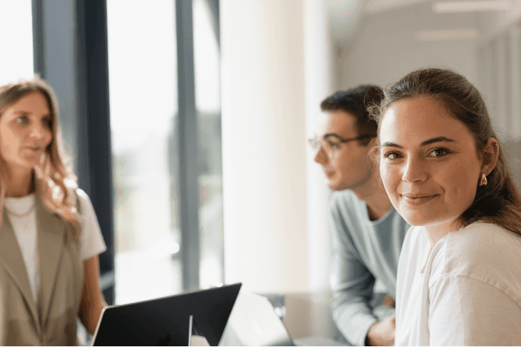 Drei junge IT-Werkstudierende sitzen an einem Besprechungstisch um einen Laptop herum