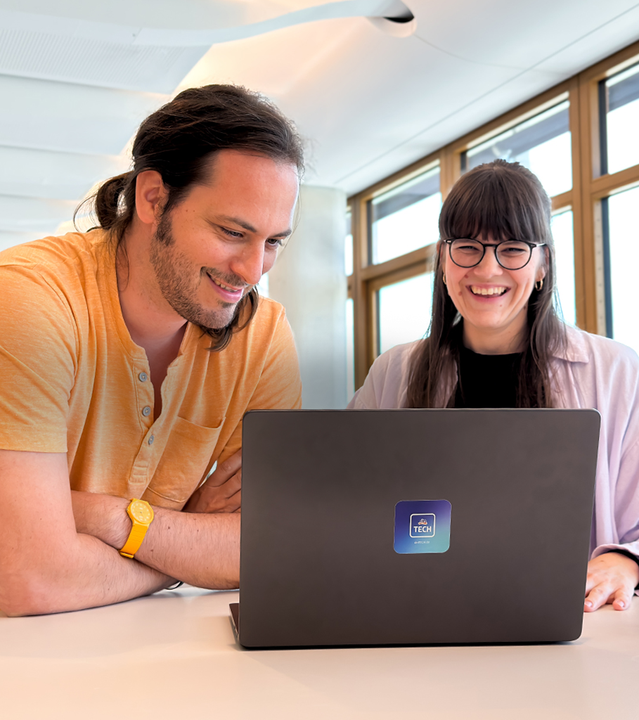 Zwei lächelnde Mitarbeitende sitzen an einem Tisch vor einem Laptop mit dmTECH-Logo. Der Mann links trägt ein orangefarbenes Hemd und eine gelbe Uhr, die Frau rechts trägt ein rosa Hemd und eine Brille. Beide schauen auf den Bildschirm und wirken engagiert und zufrieden bei der Zusammenarbeit.